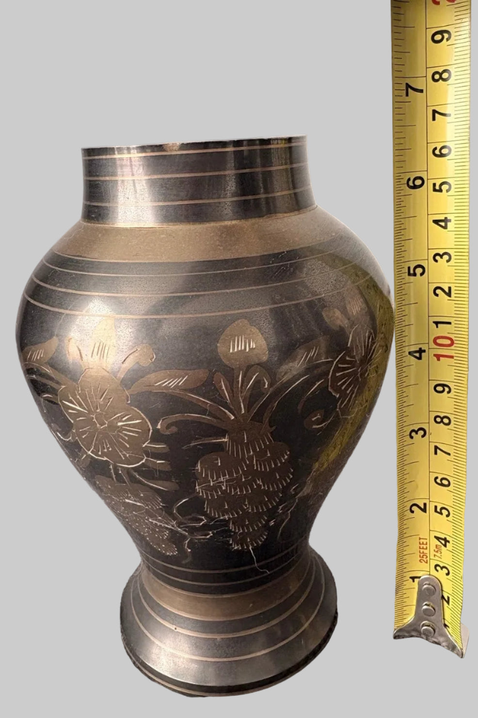 Bronze vase with floral patterns next to a tape measure, measuring 7" tall, on a grey background