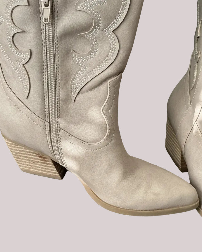 Close up of taupe cowboy boots with a small stain on the block heel, placed on a simple gray surface