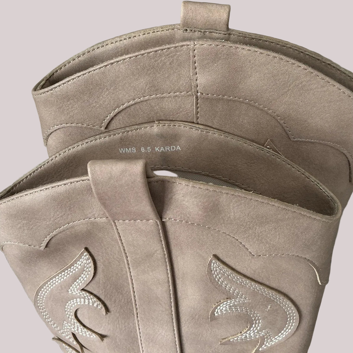 Close-up of a pair of taupe suede boots with pull tabs and intricate embroidered designs on a light gray background