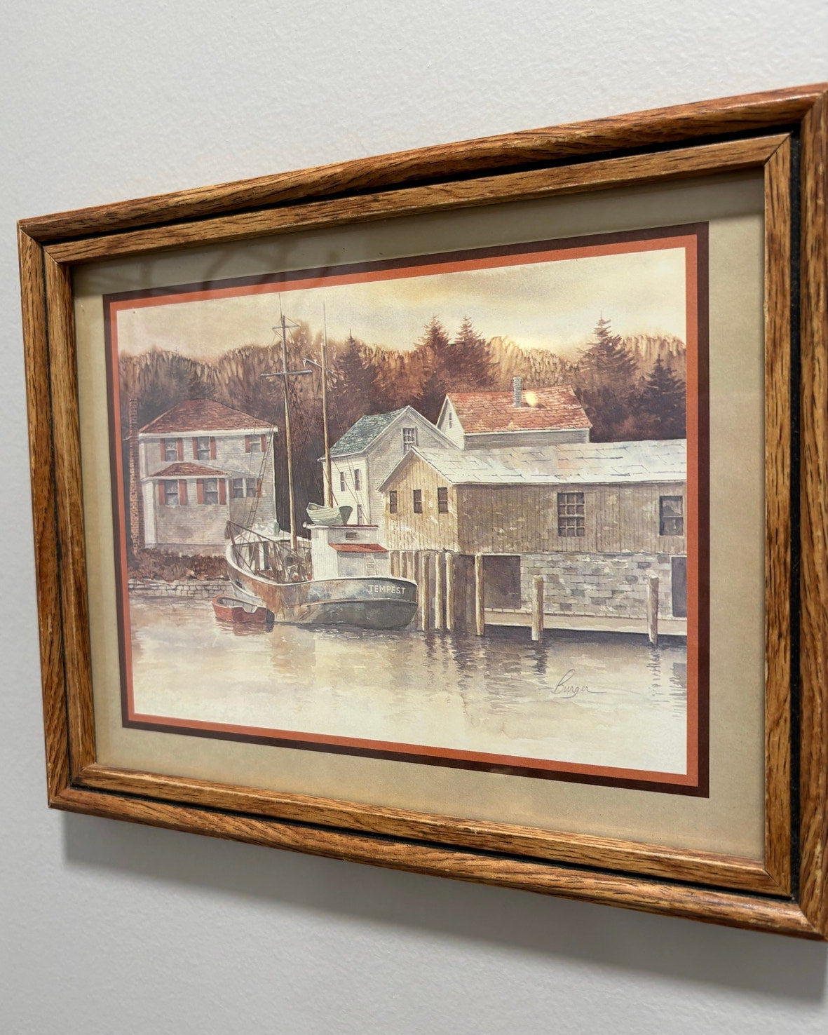 Framed painting of a coastal scene with houses and a boat on a gray wall.