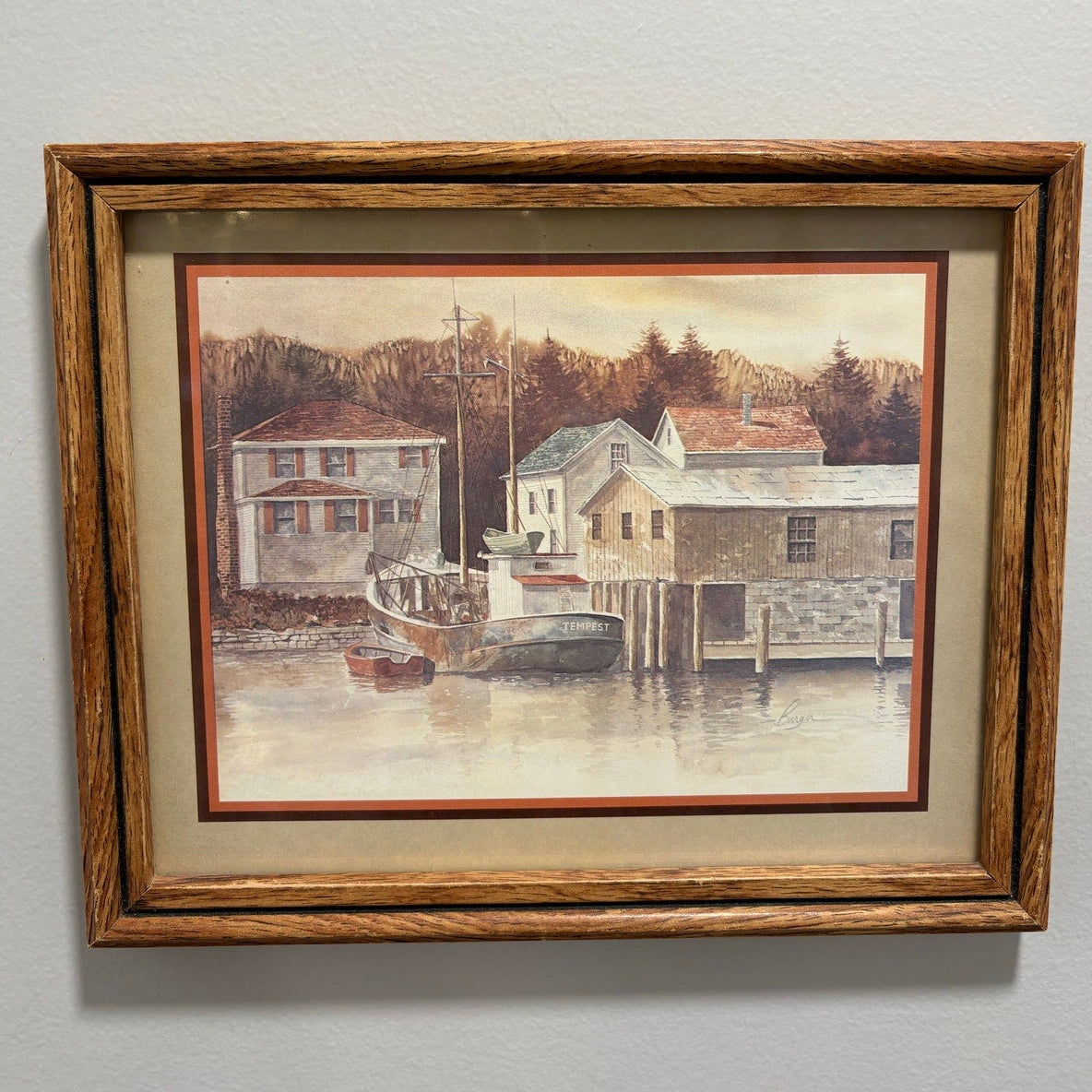 Framed painting of a coastal scene with houses and a boat on a gray wall.