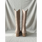 Pair of taupe knee-high boots with a block heel, on a textured linen surface.